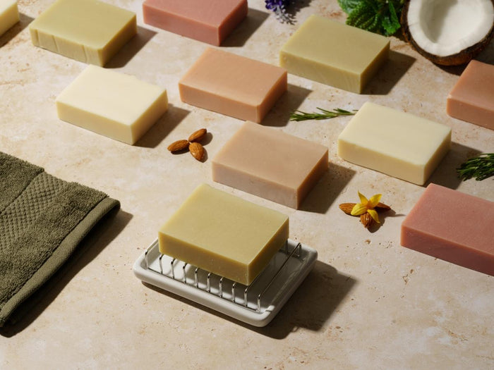 Various dry skin bars of soap on a textured surface with a green towel and next to natural soap ingredients like coconut, almonds, rosemary.