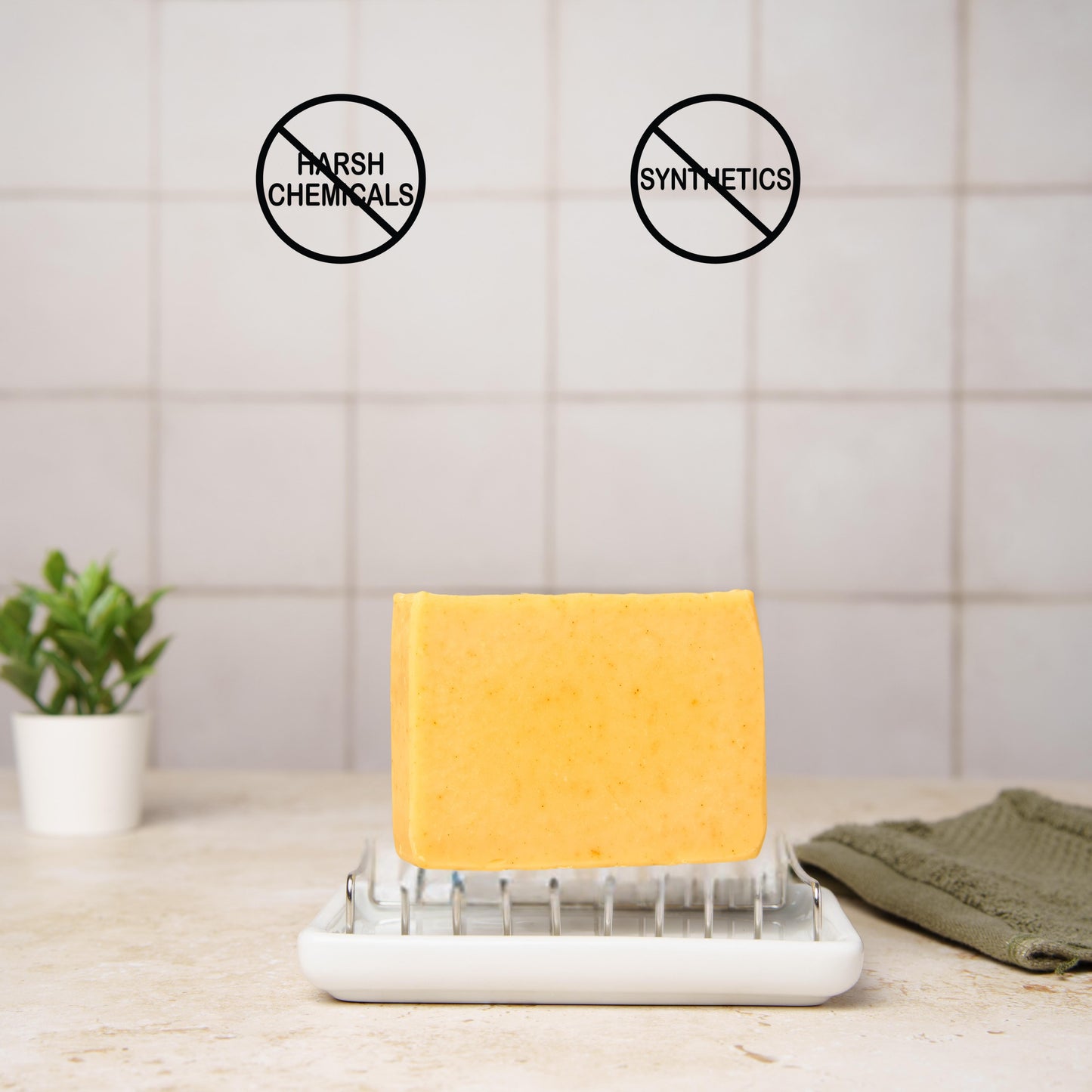 turmeric soap for sensitive skin on a soap dish with 'no harsh chemicals' and 'no synthetics' symbols above it, on a tiled bathroom wall background.